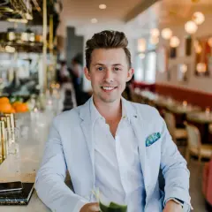 Uśmiechnięty mężczyzna w jasnoniebieskiej marynarce siedzi przy barze. Zastanawiasz się, ile zarabia manager hotelu?