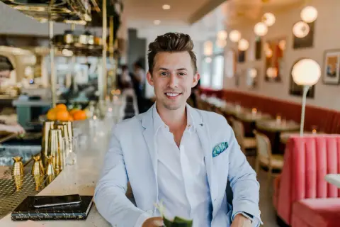 Uśmiechnięty mężczyzna w jasnoniebieskiej marynarce siedzi przy barze. Zastanawiasz się, ile zarabia manager hotelu?