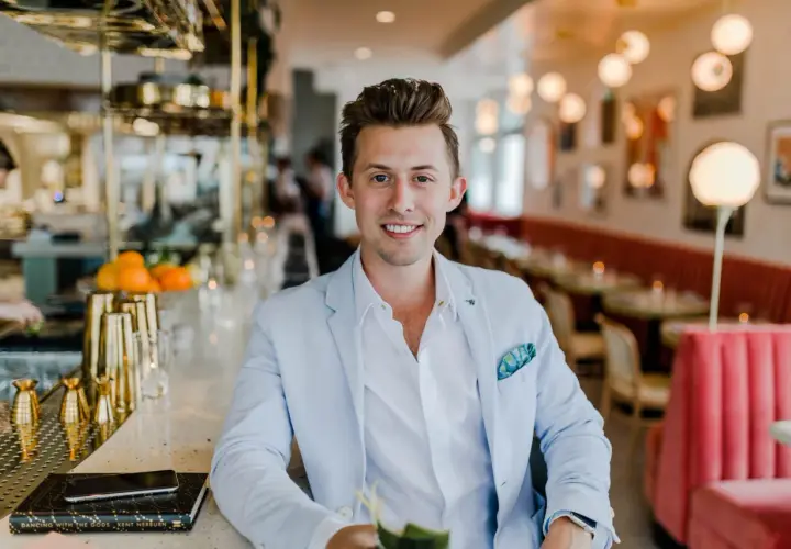 Uśmiechnięty mężczyzna w jasnoniebieskiej marynarce siedzi przy barze. Zastanawiasz się, ile zarabia manager hotelu?