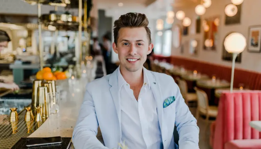 Uśmiechnięty mężczyzna w jasnoniebieskiej marynarce siedzi przy barze. Zastanawiasz się, ile zarabia manager hotelu?